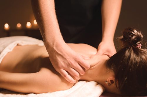 Men's hands make a therapeutic neck massage for a girl lying on a massage couch in a massage spa with dark lighting. Close-up. Dark Key.